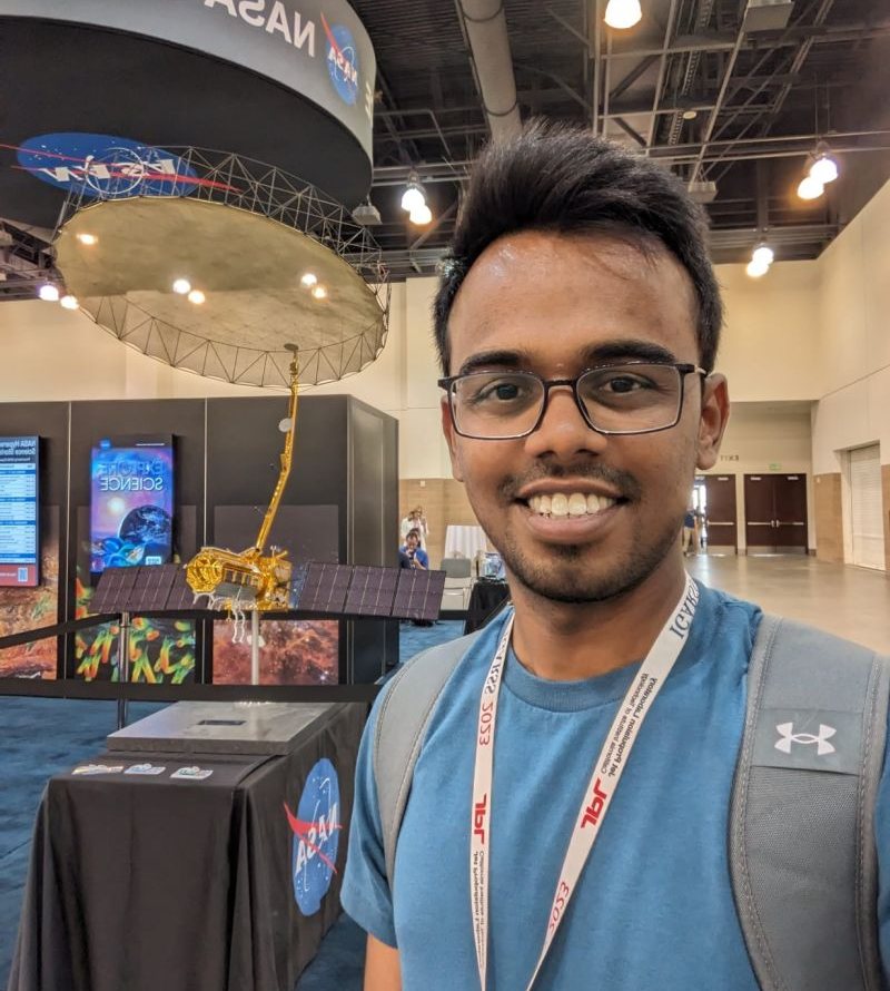 Vamshi Karanam smiling beside a gold scale model of the NISAR satellite, with its large mesh radar antenna, at the NASA exhibit.